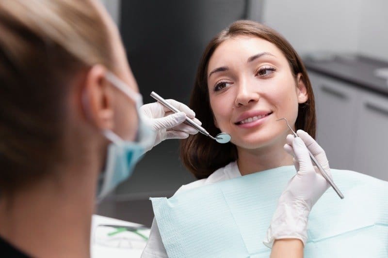 Klinik koltuğundaki kadın hastanın iki yanında ayna ve sond tutan eldivenli eller