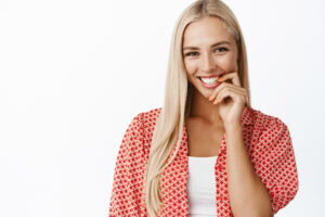 portrait of flirty feminine blond woman, biting finger and gazing coquettish at camera, smiling, standing over white background